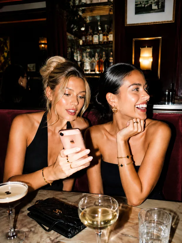 Two women laughing over cocktails, one showing the other her phone
