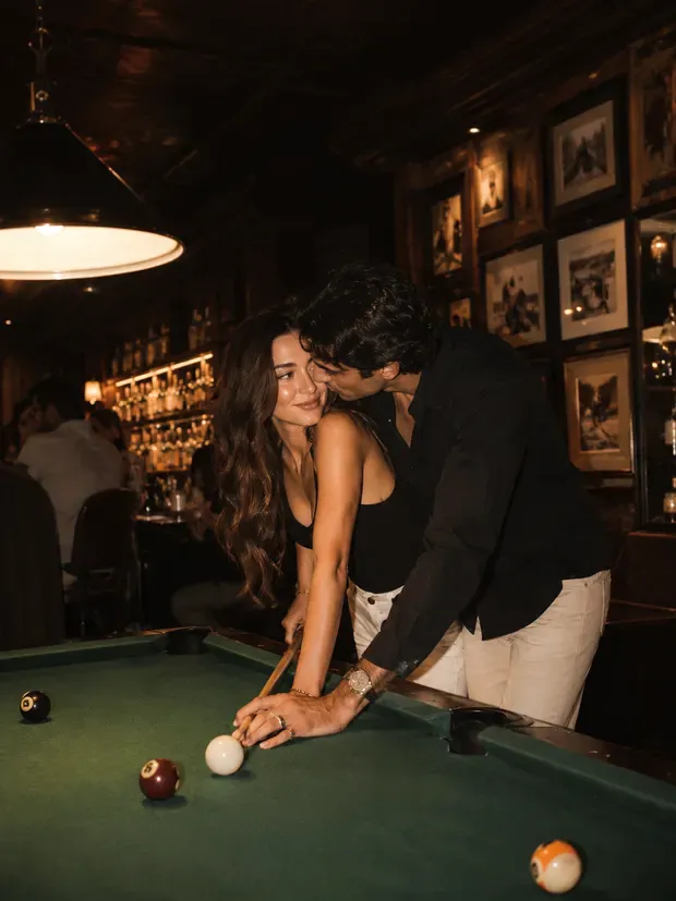 A couple leaning close over a pool table at a dimly lit speakeasy bar